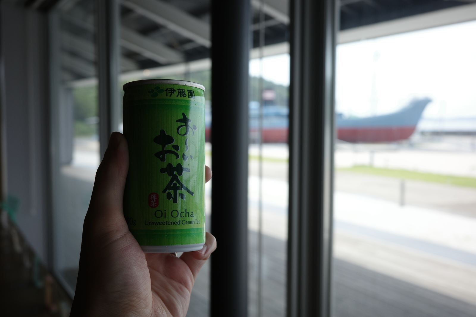 窓辺で缶入りの緑茶を持つ手。窓の外には陸に上げられた漁船が見える