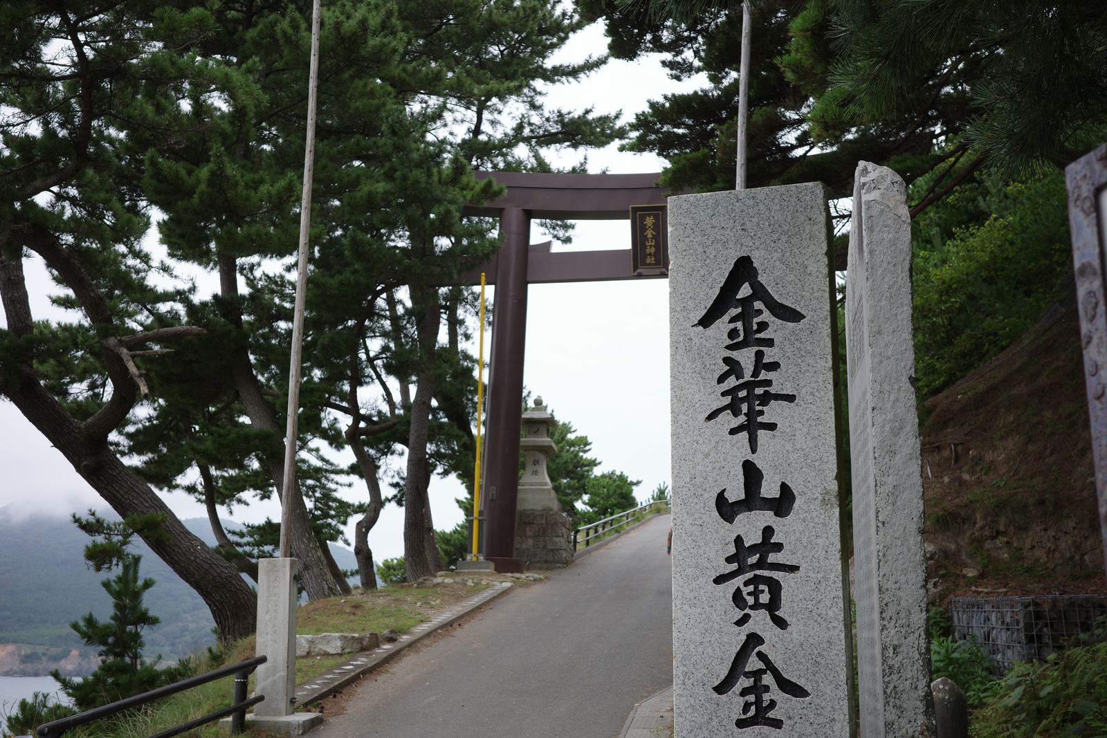 松に囲まれた坂道の先に立つ二の鳥居と、「金華山黄金…」と刻まれた石柱