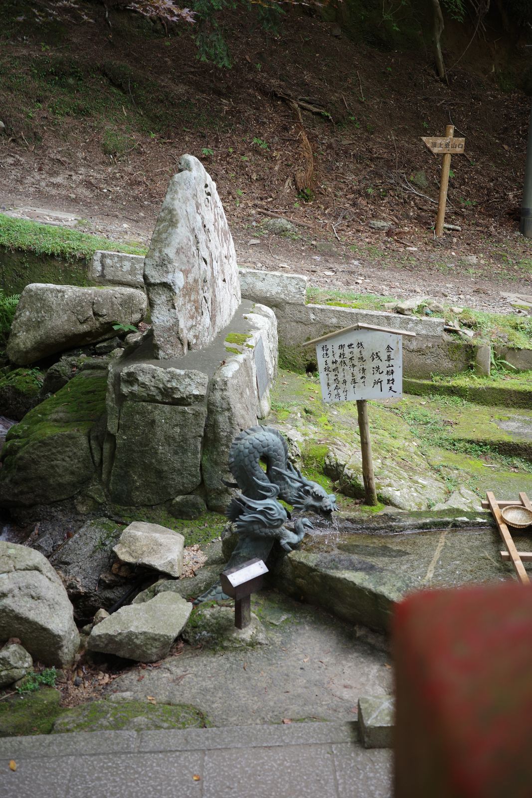 「銭洗所」と書かれた木の立て札と、口から水を注ぐ龍の像。奥に大きな石碑が立っている