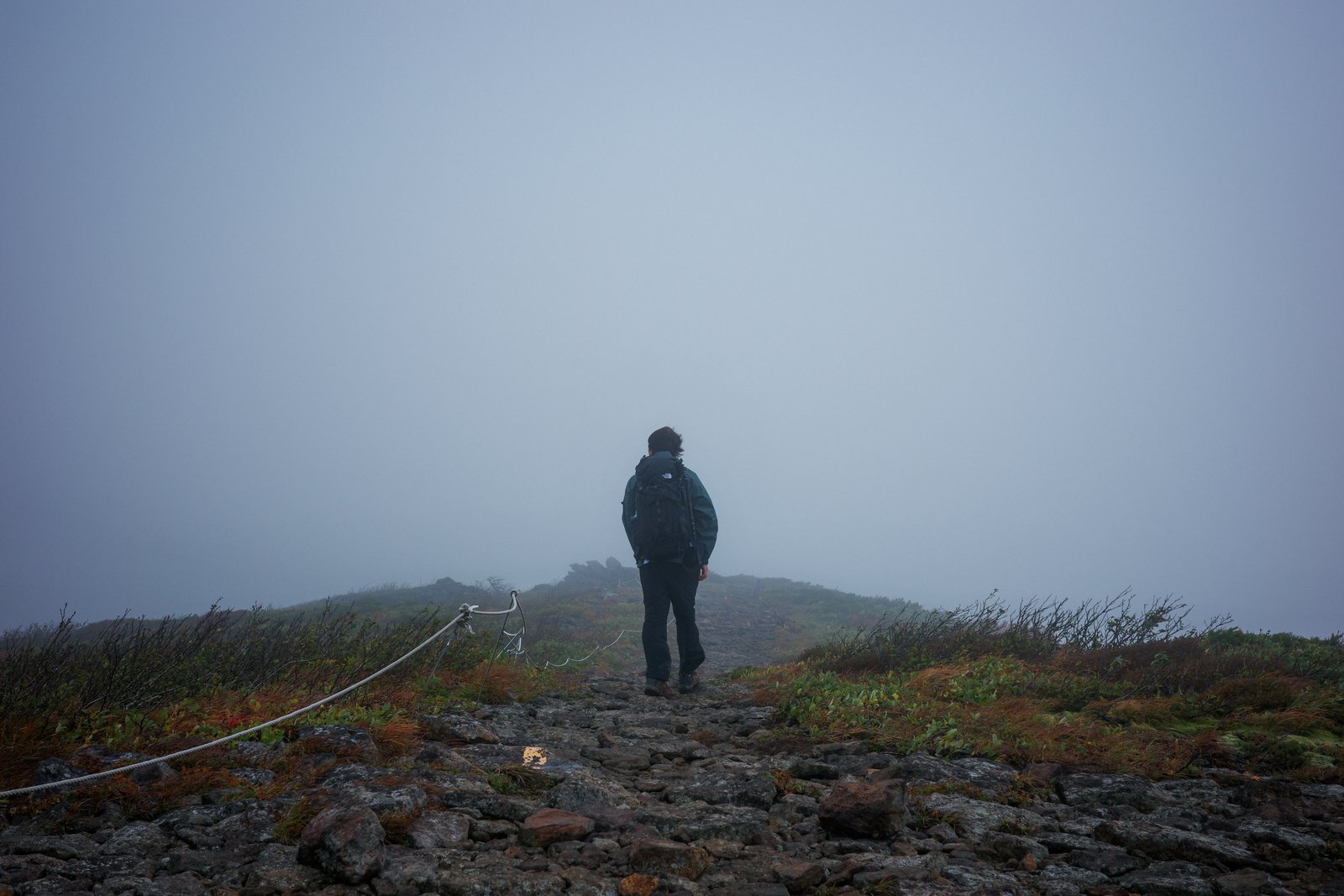濃霧に包まれた稜線の登山道に立つ、フードを被った登山者。道の脇には低い草木がある
