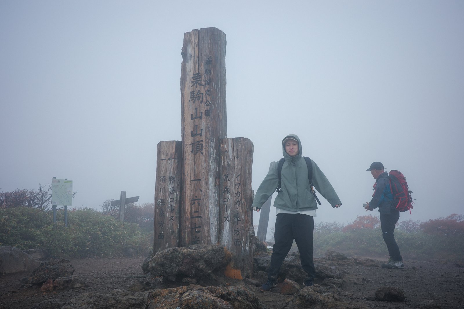 濃霧の栗駒山山頂で、フードを被ったTANAYANが木製の山頂標柱の横に立っている