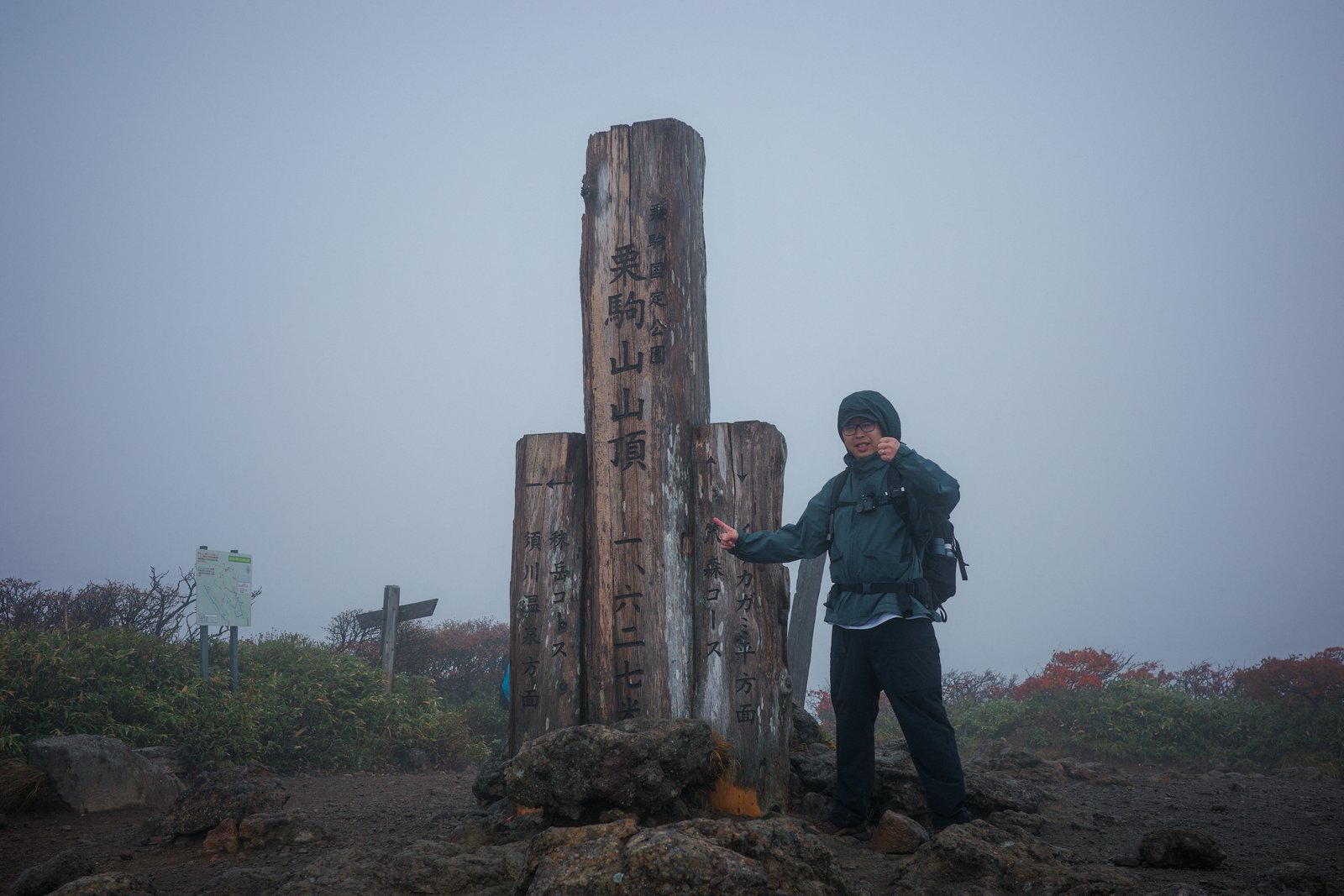 濃霧の栗駒山山頂で、山頂の木製標柱の横に立つ登山者