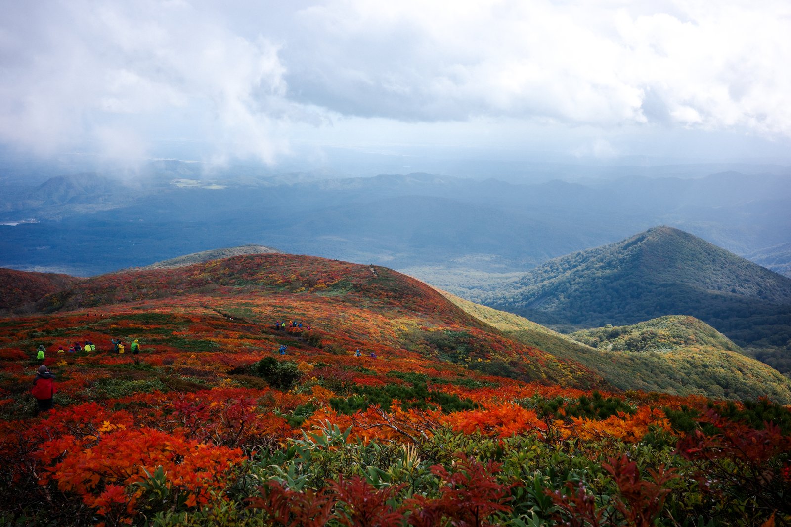 赤い紅葉に覆われた栗駒山の斜面と登山道を歩く人々。奥には山並みと雲の切れ間から光が差す平地