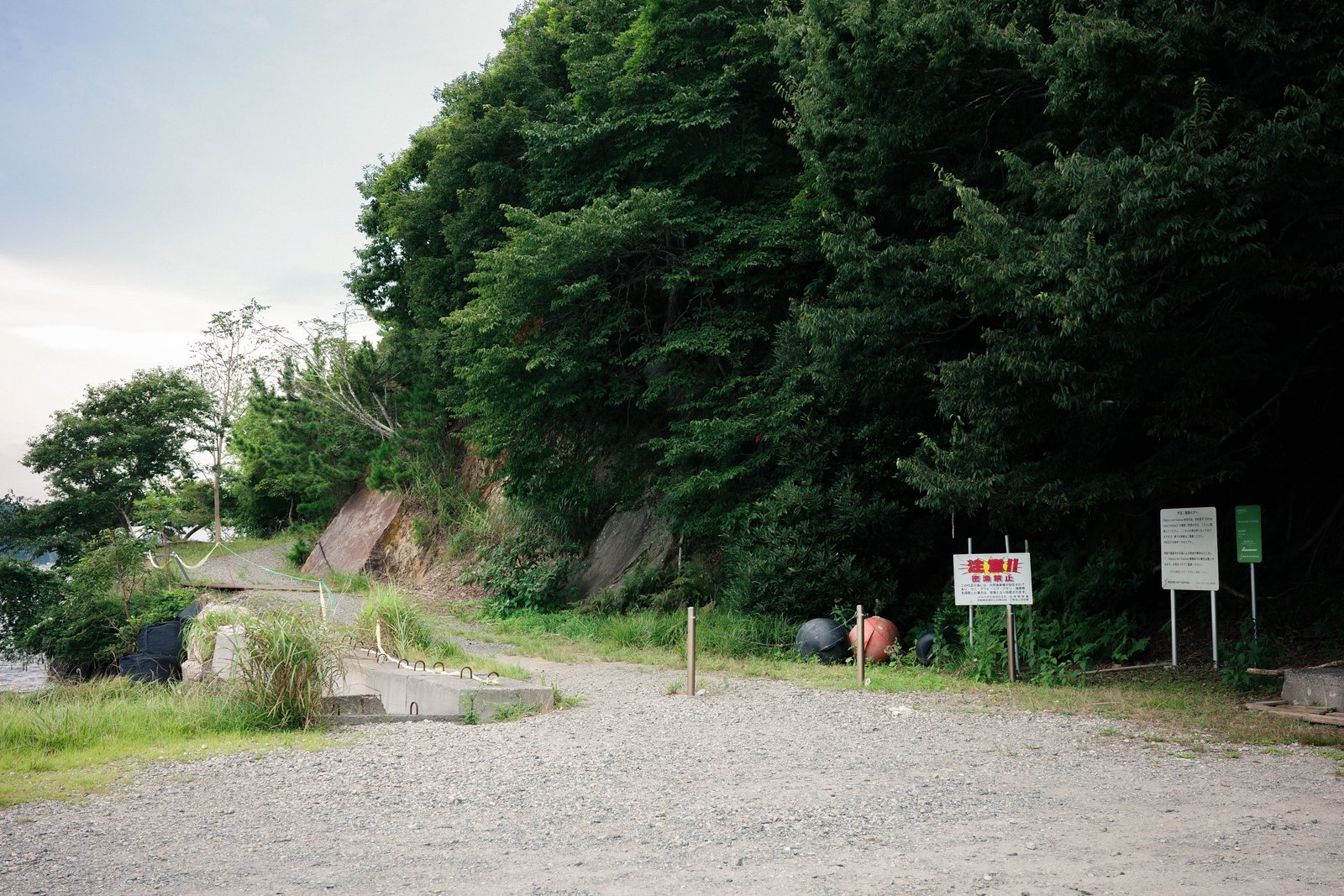 砂利敷きの広場の先に続く細い道。入口に注意書きの看板と案内板が立ち、右手は木が茂る斜面