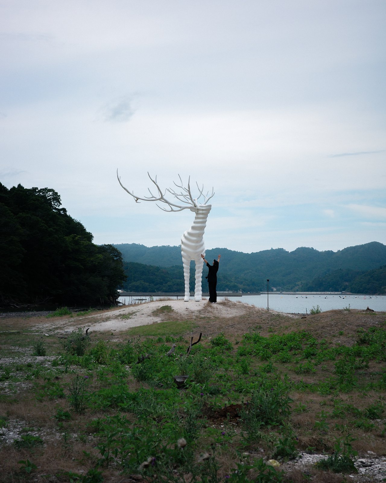小高い砂地に立つ白い鹿の彫刻と、その足元で両手を上げて立つ人物。鹿は人の何倍もの高さがある